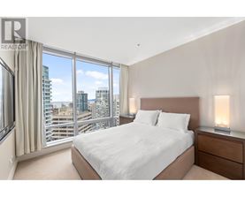 A bedroom with a large bed, a window with a view of the city, and a nightstand with a lamp.