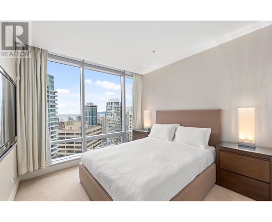 A bedroom with a large bed, a window with a view of the city, and a nightstand with a lamp.
