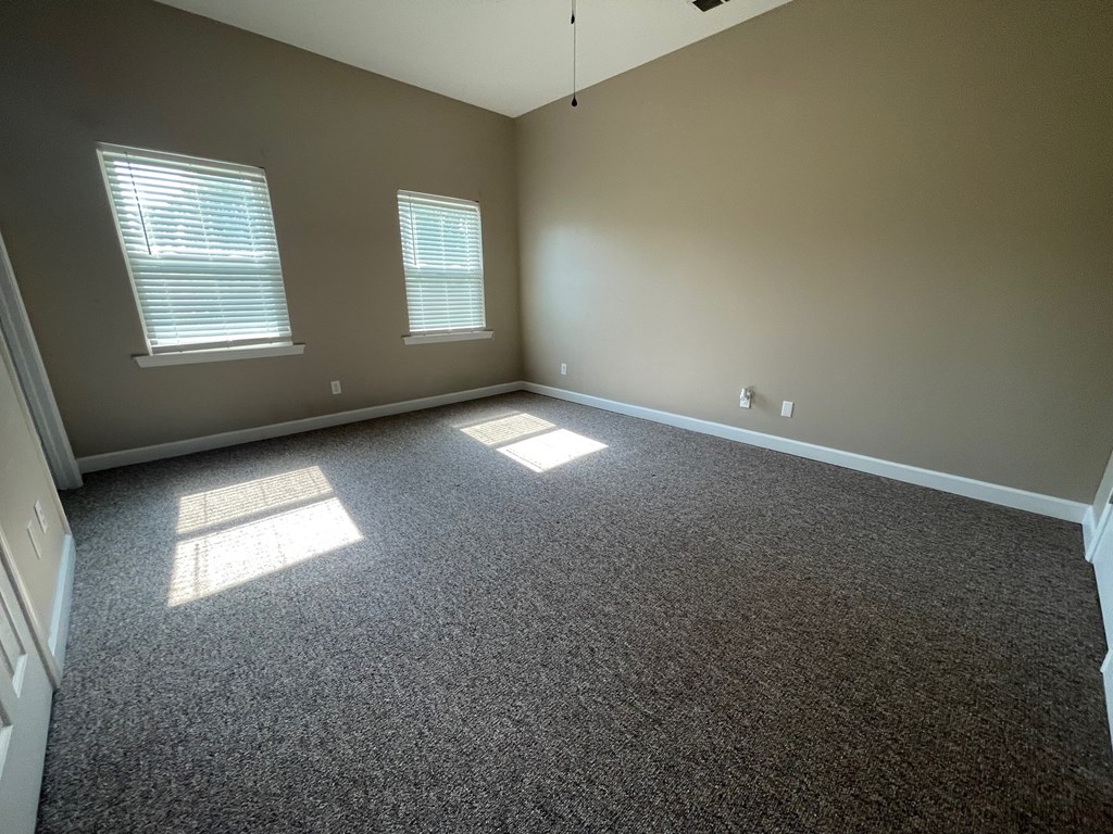 A room with grey carpet and two windows letting in sunlight.