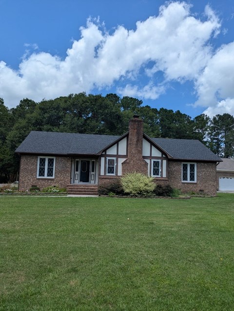 A house with a black roof and a chimney is surrounded by a grassy lawn.