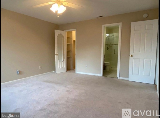 A room with beige walls and carpeting, a ceiling fan, and two doors, one of which is open.