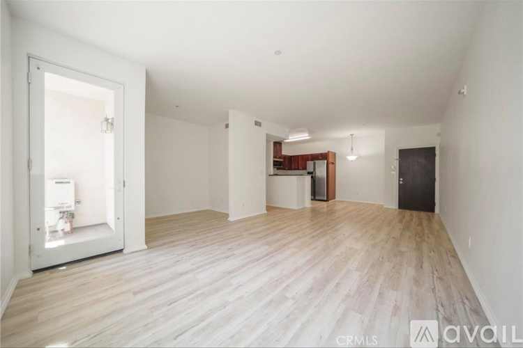 A spacious room with wooden flooring and white walls, with a door on the left and a small kitchenette in the background.