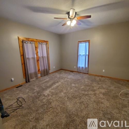 A room with a carpeted floor, a ceiling fan, and two windows with curtains.