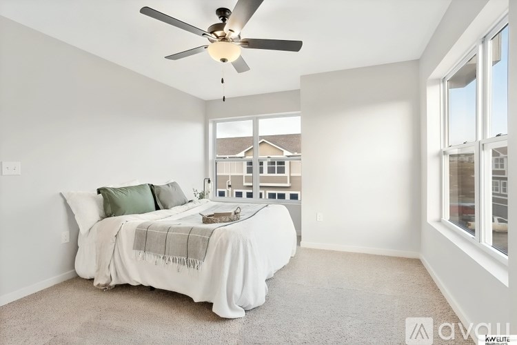 A bedroom with a bed, pillows, a blanket, a ceiling fan, and a window overlooking a building.