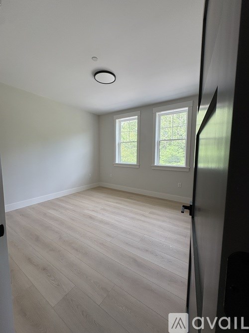 A room with a light wooden floor and a window with a view of a fence and trees.