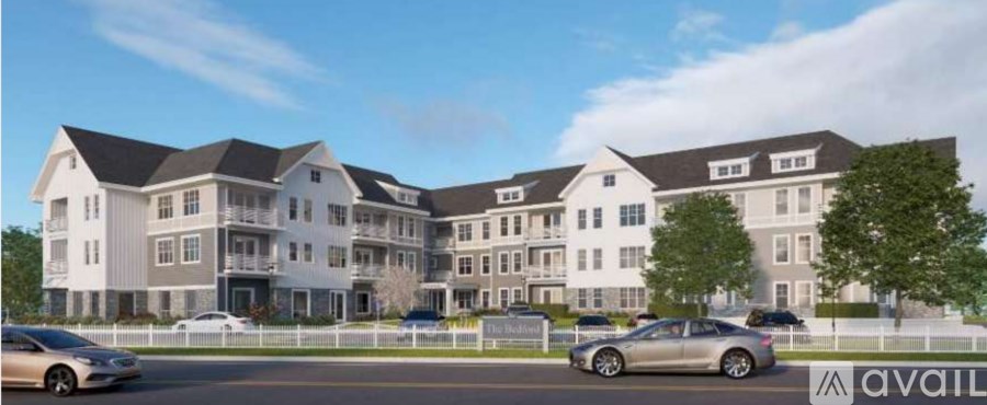 A modern residential building with multiple floors and balconies, surrounded by a white fence with cars parked in front.