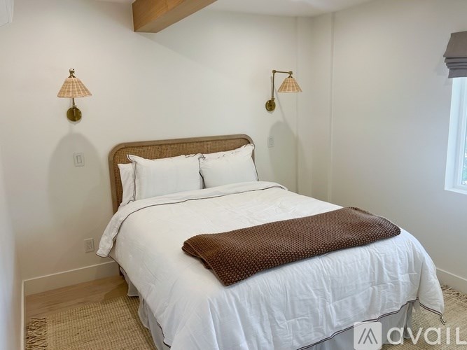 A bed with a white comforter and a brown throw is in a room with two wall lamps.