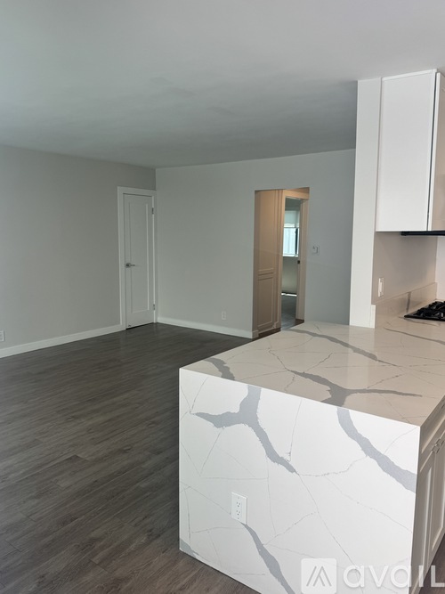 A white kitchen island with a marble top is in the foreground of an empty room with wood flooring and white walls.