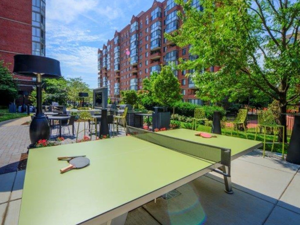 A ping pong table is in the foreground of a sunny outdoor area with trees and buildings in the background.
