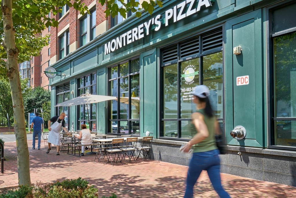 A woman in a green tank top and blue jeans is walking past a pizza restaurant.