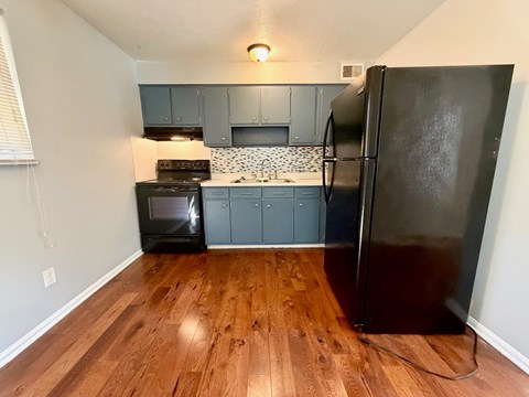 A black refrigerator stands in a kitchen with wooden floors and grey cabinets.