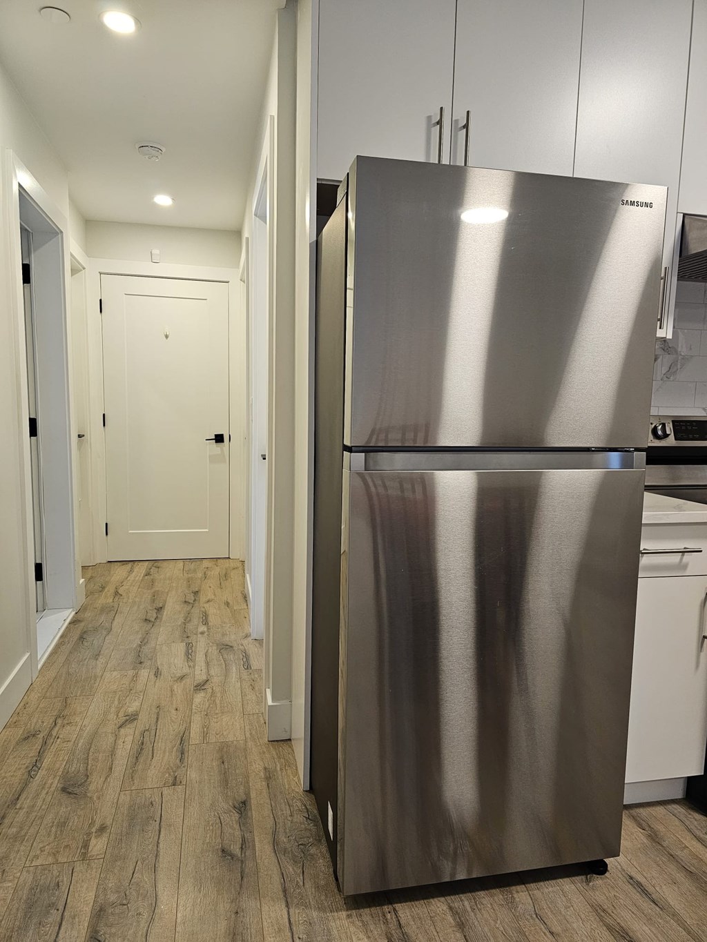 A stainless steel refrigerator stands in a kitchen with wood flooring.