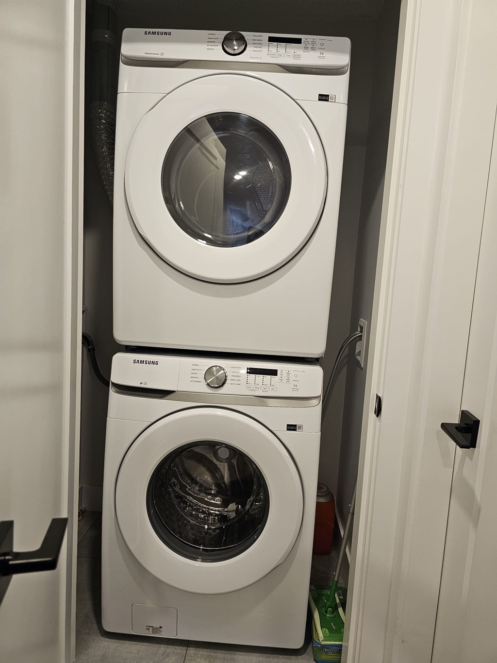 Two white Samsung front load washing machines stacked on top of each other in a small laundry room.