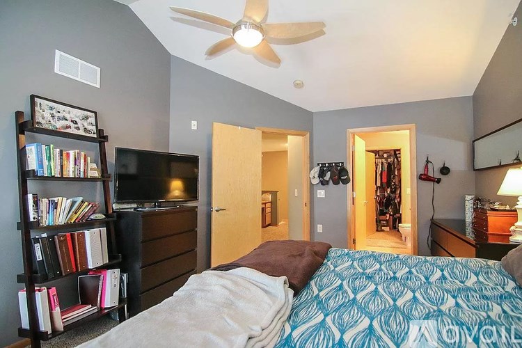 A bedroom with a bed, a bookshelf, a ceiling fan, and a doorway leading to another room.