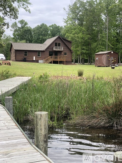 A wooden dock leads to a body of water with a house in the background.