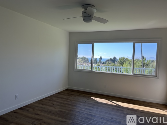 A room with a ceiling fan and a large window overlooking a fence and trees.