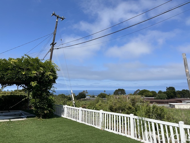 A white fence in front of a green lawn with a view of the ocean in the distance.