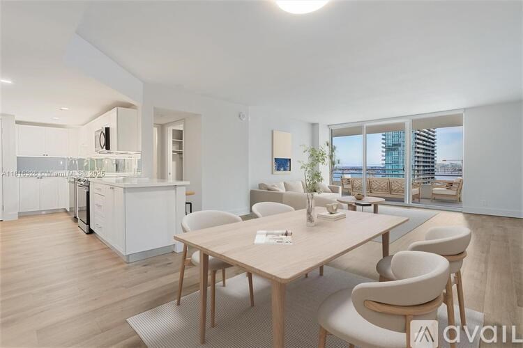 A modern kitchen with a dining table and chairs in the foreground and a balcony with a view of the city in the background.