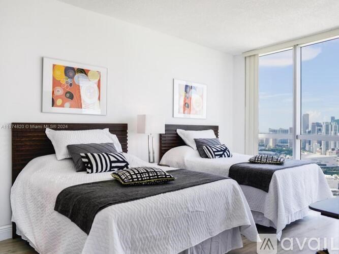 Two twin beds with white bedspreads and black pillows in a room with a large window overlooking a cityscape.