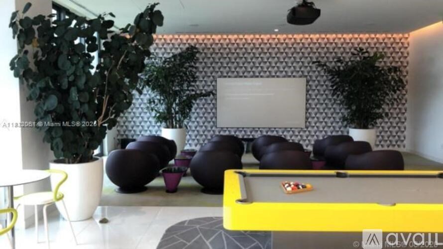 A pool table with a yellow cloth and a white cue ball is in the foreground of a room with a patterned wall and potted plants.