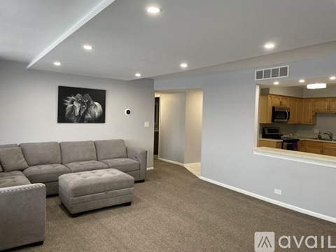 A living room with a grey sofa and ottoman, a painting on the wall, and a kitchen area in the background.