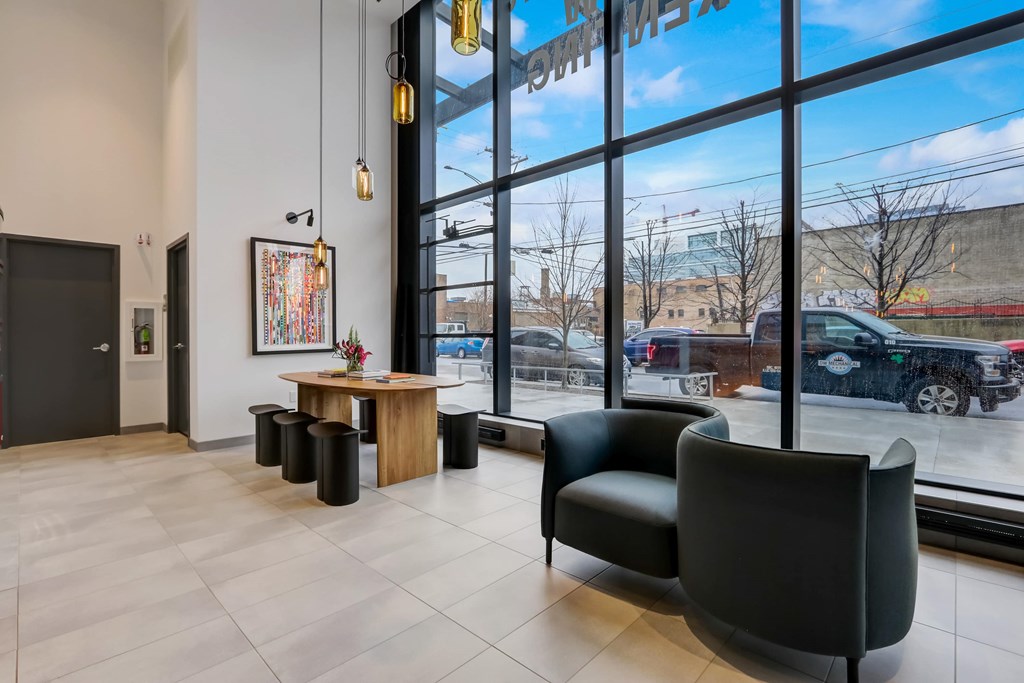 A black chair sits in front of a large window in a room with a wooden table and a painting.