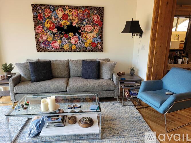 A living room with a grey sofa, a blue chair, a glass coffee table and a large floral tapestry on the wall.
