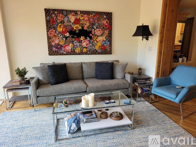 A living room with a grey sofa, a glass coffee table, a blue chair, and a large floral tapestry hanging on the wall.