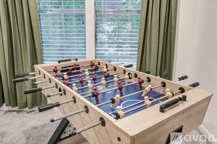 A wooden foosball table with red and black players is in front of a window with green curtains.