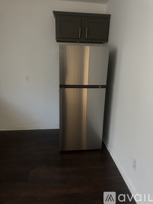 A stainless steel refrigerator with black handles and a black top sits in a room with a wooden floor.