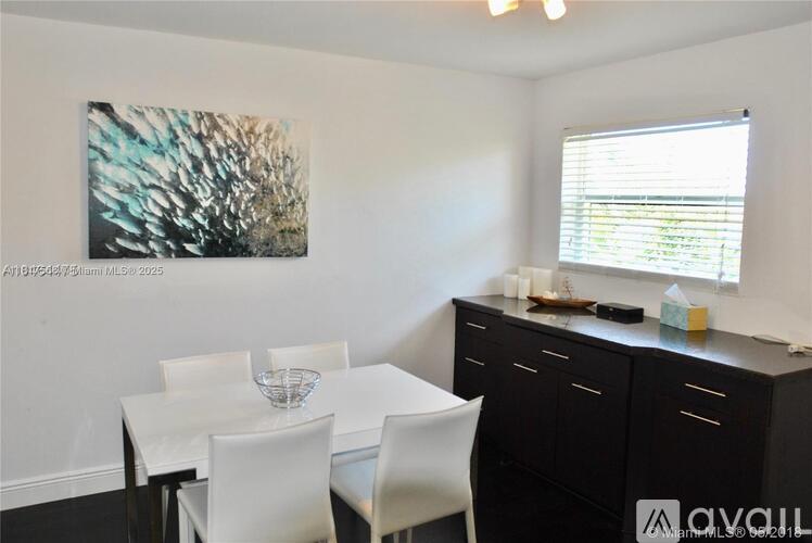 A dining room with a white table and chairs, a painting on the wall, and a window with blinds.