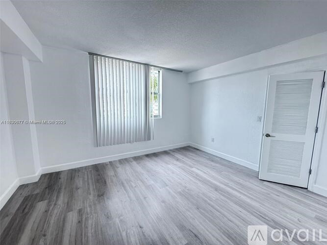 A room with wooden flooring and white walls, with a window covered by vertical blinds.