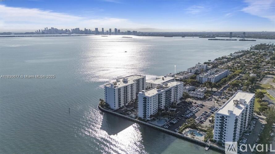 A large white building sits on the edge of a body of water with a city skyline in the background.