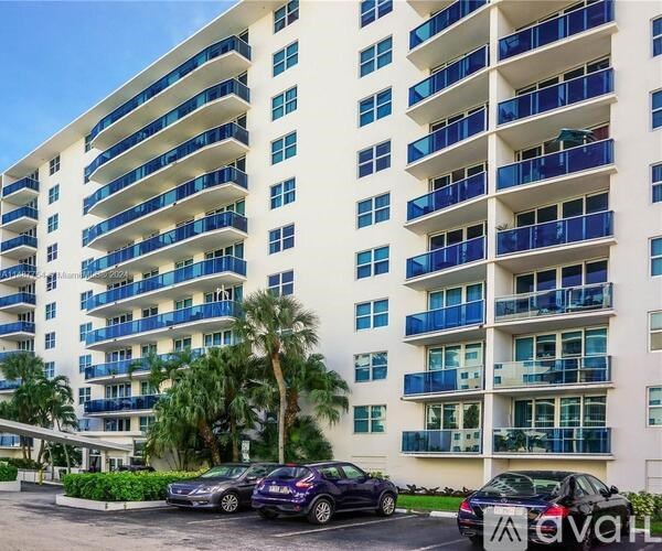 A tall white building with blue balconies and cars parked in front.