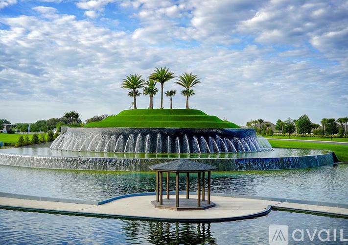 A gazebo sits on a pond in front of a waterfall and palm trees.
