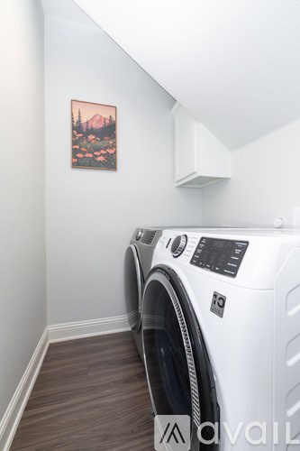 A washing machine sits in a laundry room with a painting of a forest scene on the wall.