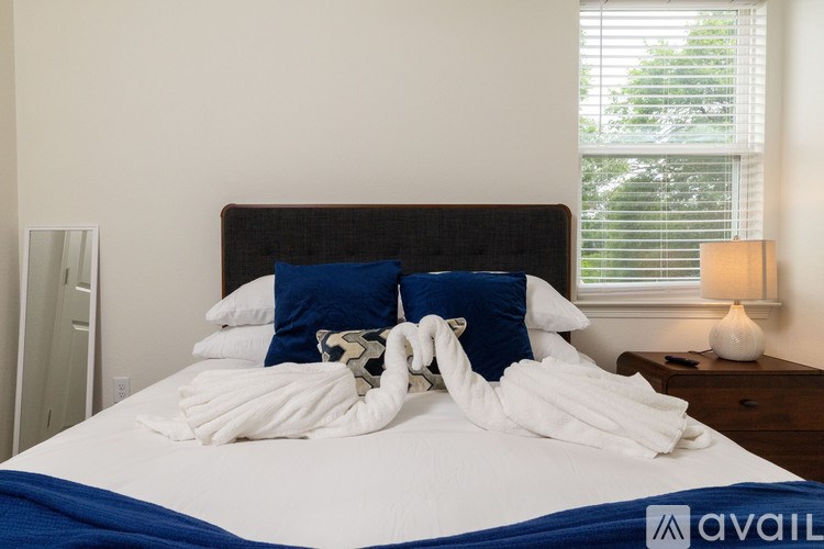 A bed with a white comforter and a blue blanket is in the foreground with a window in the background.