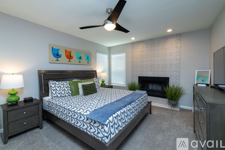 A bedroom with a patterned bedspread, a ceiling fan, and a fireplace.