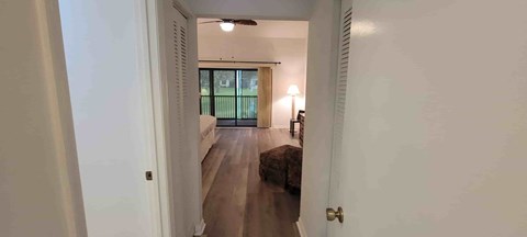A hallway with a door on the left and a bedroom with a bed and a lamp on the right.