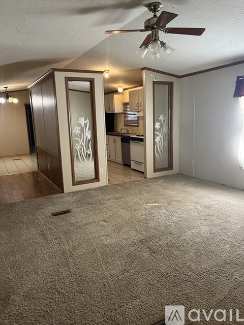 A carpeted room with a ceiling fan and a kitchen area with cabinets and a counter.