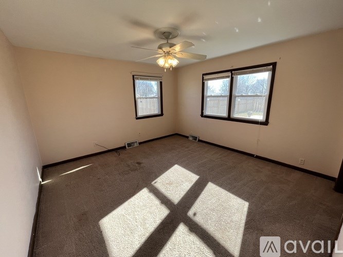 A room with a carpet, a ceiling fan, and two windows.