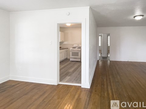 A room with wooden floors and white walls, leading to a kitchen area.
