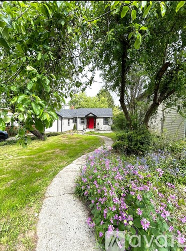 A small house with a red door is surrounded by greenery and purple flowers.