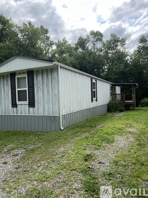 A white mobile home with a brown door and a window is surrounded by grass and trees.