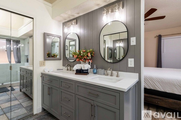 A bathroom with a vanity, two mirrors, and a bed in the background.