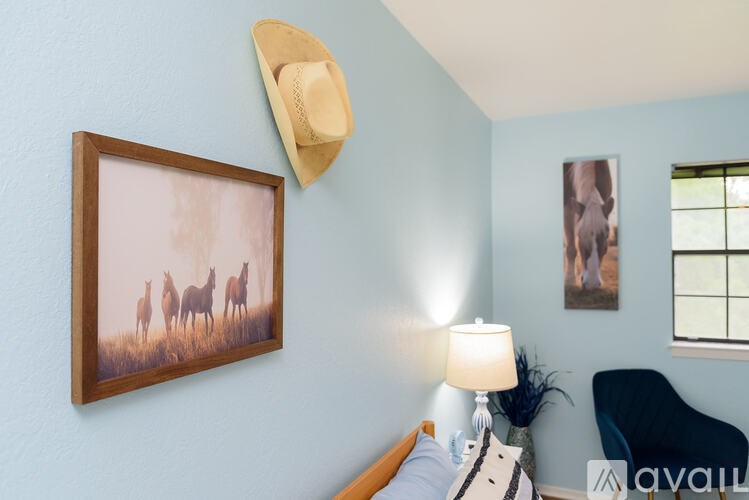 A bedroom with a bed, a lamp, a painting of horses, a hat, and a chair.