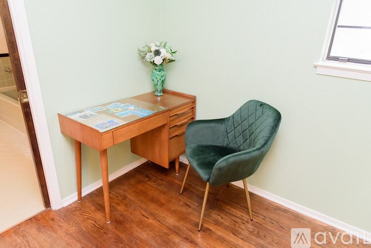 A chair with a green seat and wooden legs sits next to a desk with a vase of flowers on it.
