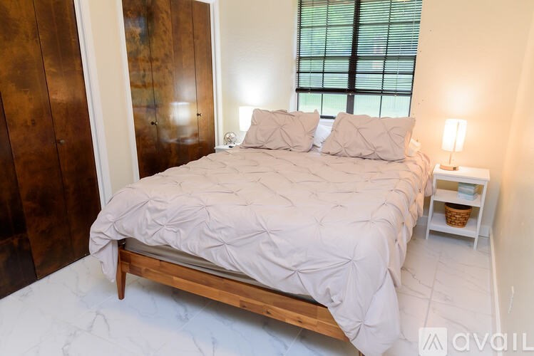 A bed with a white comforter and two pillows sits in a room with a wooden headboard and a window with blinds.