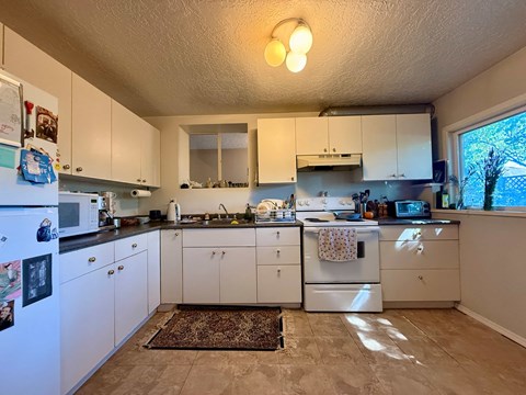 A kitchen with white cabinets and appliances, a rug on the floor, and a window letting in natural light.