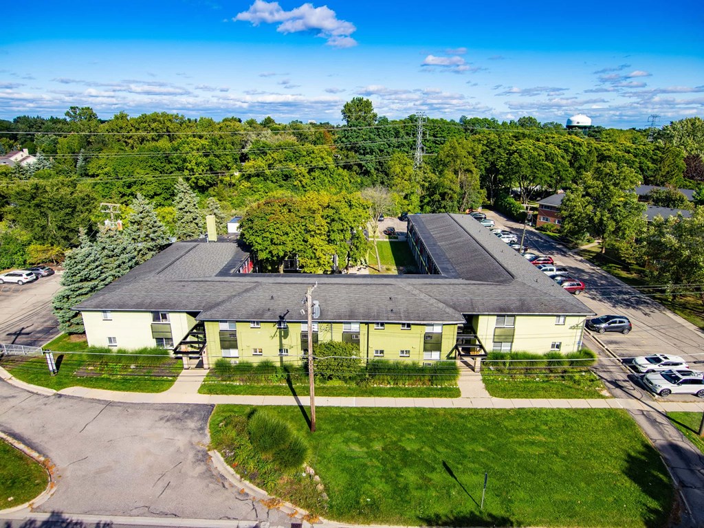 A large green building with a grey roof is surrounded by a grassy area and trees.